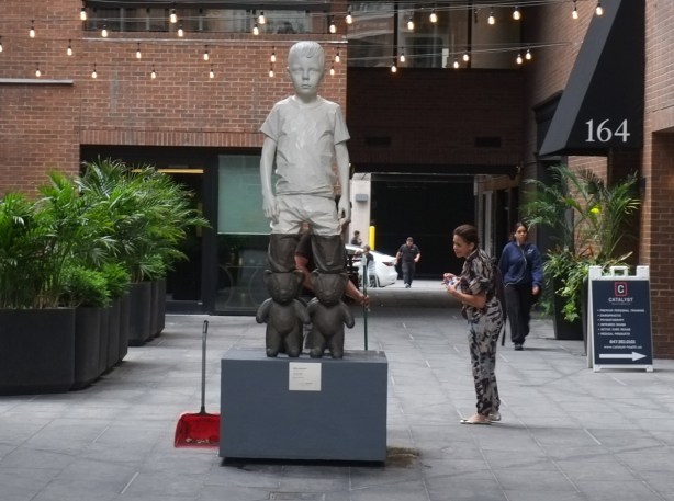 in courtyard at 80 cumberland, a statue by Italian artist Willy Verginer, a boy all dressed in white is kneeling on 4 grey teddy bears