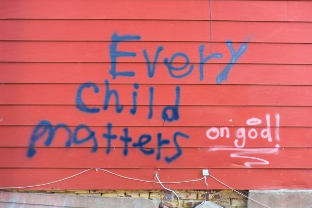 on a red wood garage wall, in blue spray paint, text that says every child matters, on god