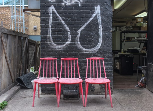 three red chairs, outside, by a black wall with white drawing that is either angel wings or eyes,