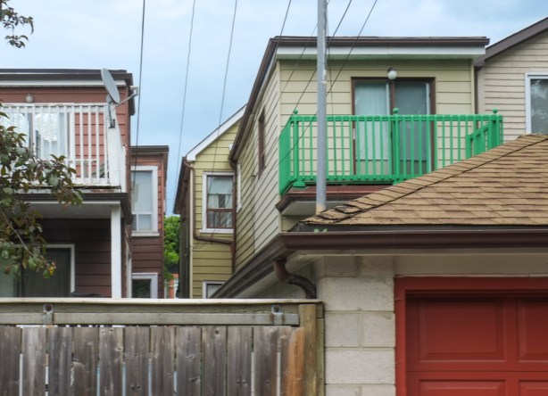 in an alley, view of back of houses, different depth of buildings, wood fence, cement block garage with red door, balcony with bright green railing, 