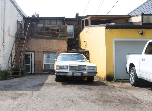 old Mercury Grand Marquis car from the early 1990s, pale yellow, parked in alley beside yellow garage