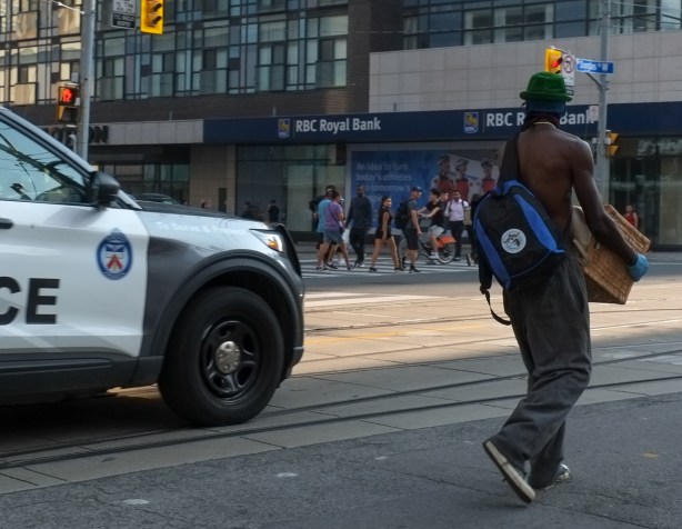 a topless young man wearing a hat and jeans, and carrying a bag over his shoulder, in middle street, police car passes by, people on sidewalk turn to look because police car has short blast of the siren