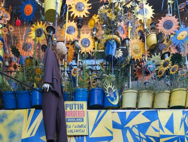 on a balcony in Kensington, many sunflowers, both real and fake, plants in blue and yellow pots, an effigy of putin, and a sign that says putin murdered over 200,000 men women and children