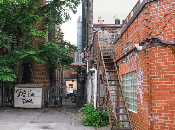alley view, metal exterior stairs, window in red brick building, old brick smokestack, balconies on upper level, archway over lane to exit to street