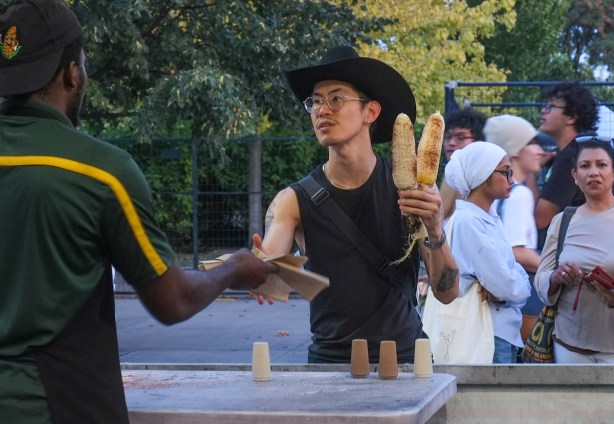 man wearing black cowboy hat is buying two cobs of corn to eat