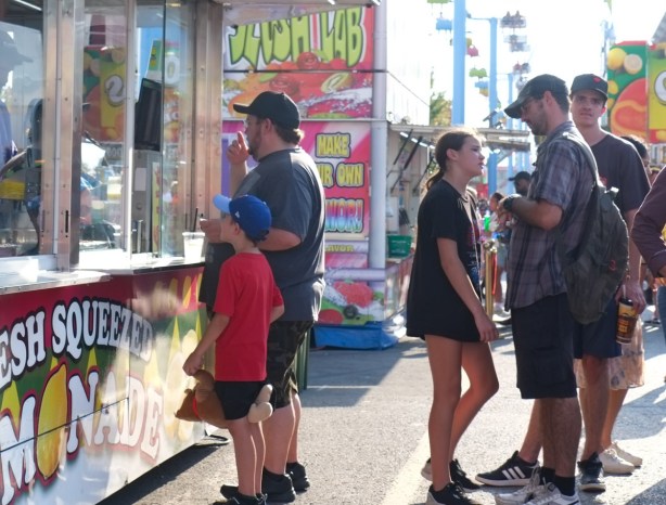 at a food stand, lemonade, at the ex, one father is buying food for his son, another father is arguing with his daughter