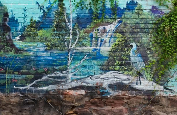 mural in cottage lane by b c johnson, a heron stands on a fallen log on a rock on shoreline, birch tree nearby, water, and a waterfall in the background