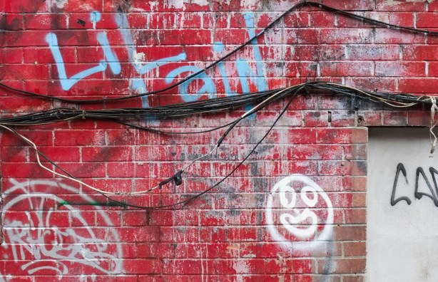 exterior brick wall, painted bright red, with many black wires in a bundle crossing it,, white graffiti happy face, other blue graffiti text lil 