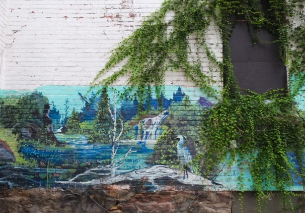 on a wall by a black door, overgrown with ivy, door looks like tree trunk, also mural in cottage lane by b c johnson, a heron stands on a fallen log on a rock on shoreline, birch tree nearby, water, and a waterfall in the background