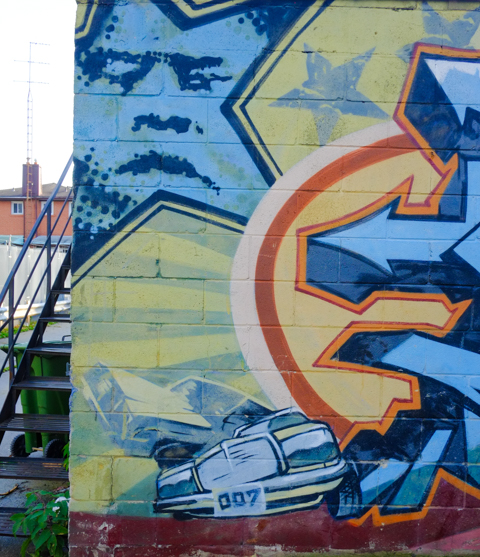 close up of a painting on exterior of garage in an alley, blue star with a man's face in it, front end of car with licence plate 007