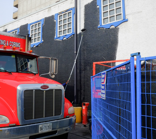 truck with red cab is parked beside a white building that is being painted black, person with a paint roller on a very long pole