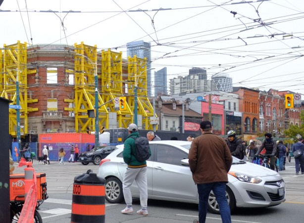a white car partially blocks the intersection as it goes southbound on spadina, pedestrians have to go around it while crossing on their green light. northeast corner of queen and spadina in the background