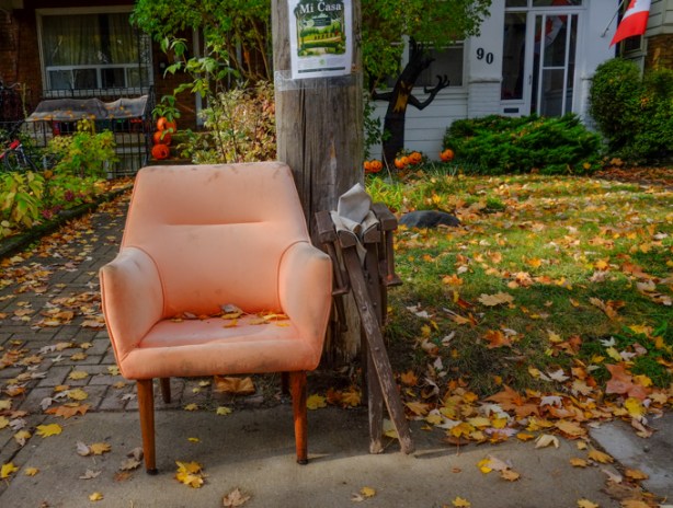 a pale orange, faded, fabric covered armchair sits by the curb on a sidewalk in front of a house, fallen autumn leaves on the ground