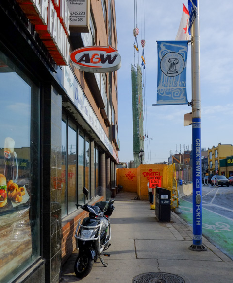 sidewalk in front of A & W, motorcycle parked there, sidewalk ends with orange fence because of construction, utility pole by sidewalk is wrapped in blue with Greektown Danforth written on it, also a blue and white Greektown banner on pole as well as Canadian flag