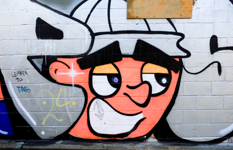 big orange cartoon face, with white cap, mural on a garage in an alley