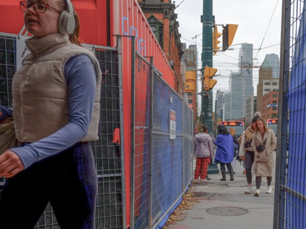 a woman wearing headphones walks past a construction site, on Spadina, other people on sidewalk crossing at traffic lights