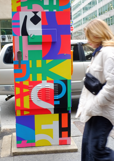 woman walking past pa metal box on sidewalk, traffic signal box, that has been covered with street art painting of abstract letters of the alphabet