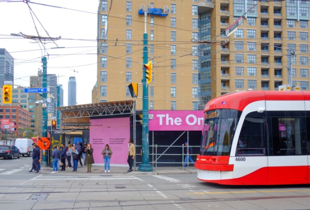 TTC streetcar on Queen, waits at red light at Spadina, people on the corner, tall building behind, pink signs advertising ontario line. 