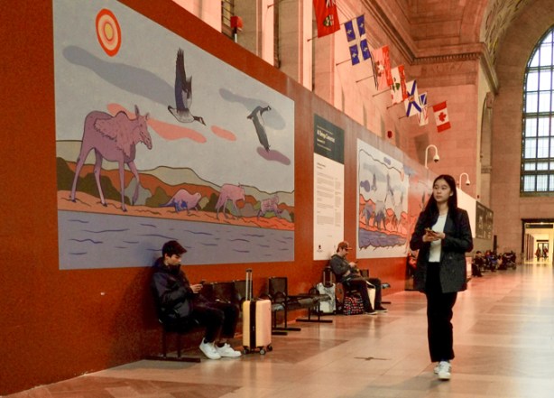 a young woman walks past large artworks on the walls of the main hall in union station, title of art is All beings connected and the artist is August Swinson