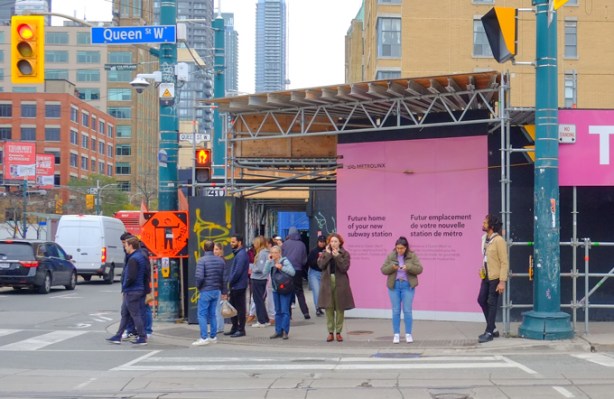 people waiting on the southwest corner of padina and queen, waiting for their green light, pink hoardings for metrolinx construction behind them