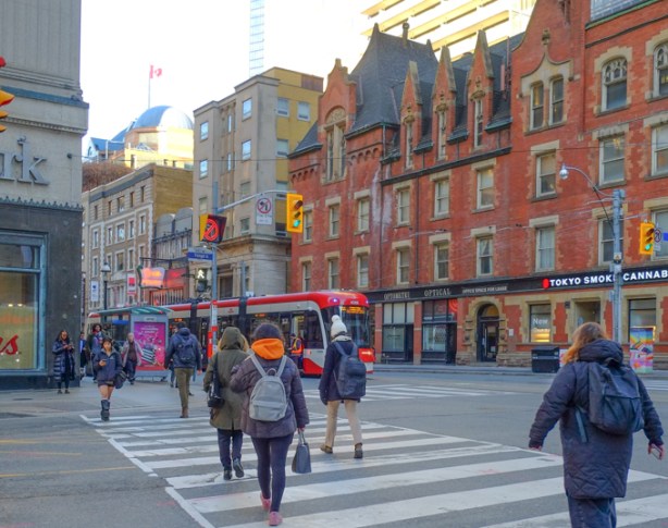 people crossing intersection of college and yonge, walking westward, towards TTC streetcar that is heading east, in front of College Park, with old red brick building on the north side of college street