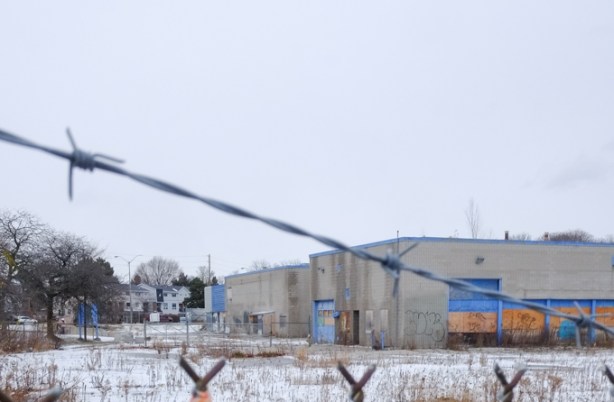 chainlink fence with strands of barbed wire on top, with empty old building and snowy covered vacant lot behind