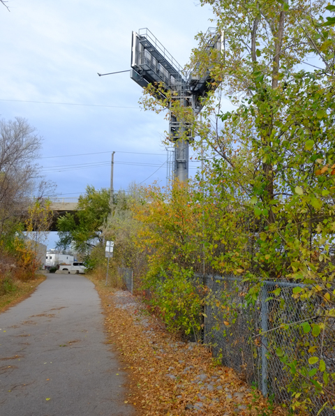 looking north up don mills trail towards york mills road, where there is a bridge over the railway tracks, a large pair of billboards on a tall pillar, dead leaves beside path, shrubs and weeds along the fence