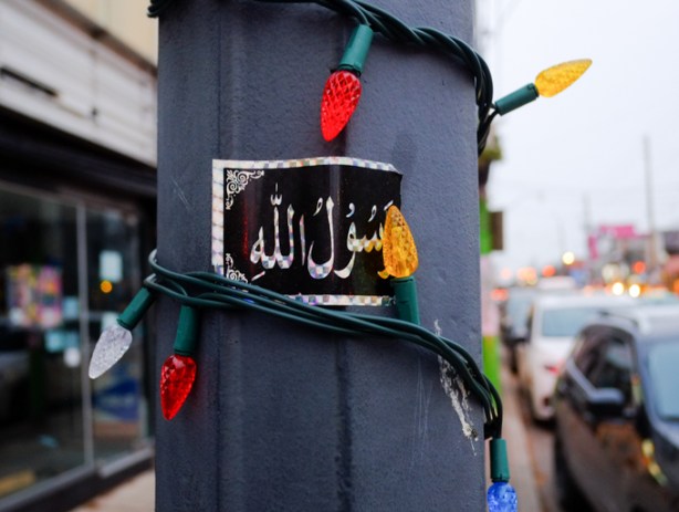 sticker with arabic writing on it, on a utility pole that is wrapped in christmas lights 