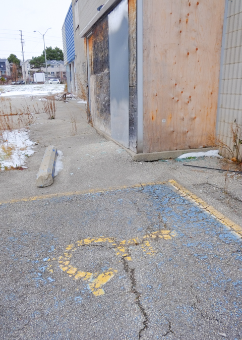 cracked pavement and a faded yellow wheelchair symbol in front of a derelict building waiting to be demolished, once was a car dealership