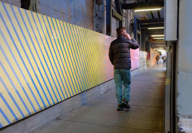man talking on his phone, walks under covered scaffolding, beside hoardings with painted diagonal stripes in yellow and blue