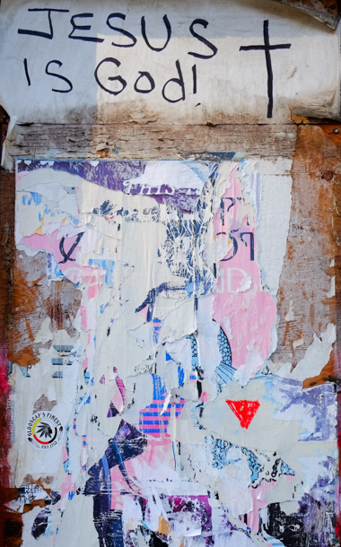 layers of ripped graffiti paste ups and posters on wall of an abandoned building with graffiti text written across the top that says Jesus is God