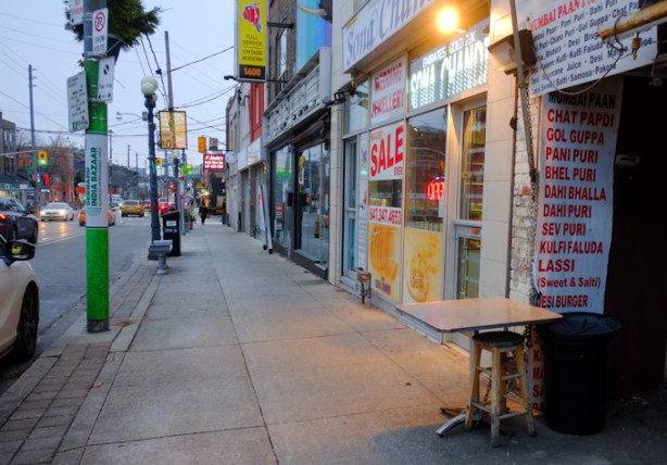 small metal table on sidewalk outside restaurant, list of food on wall, like lassi, sev puri, panu puri, and gol guppa, evening, street scene