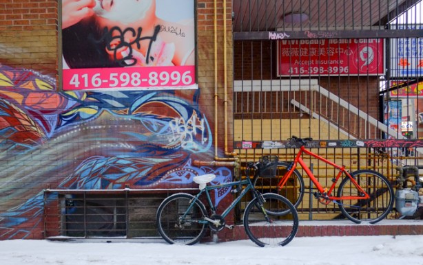 bikes chained to fence, in snowy alley, with street art on the wall and an ad too