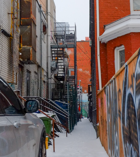 looking down the space between two houses, graffiti on the fence, snow on the ground, multiple exterior staircases