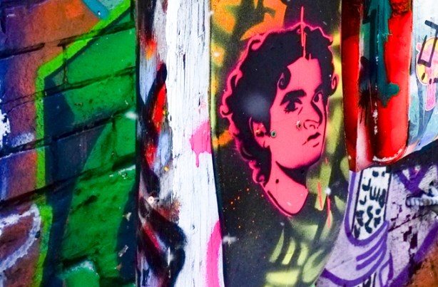 pink man's portrait painted on half of a skateboard, attached to a pole in graffiti alley