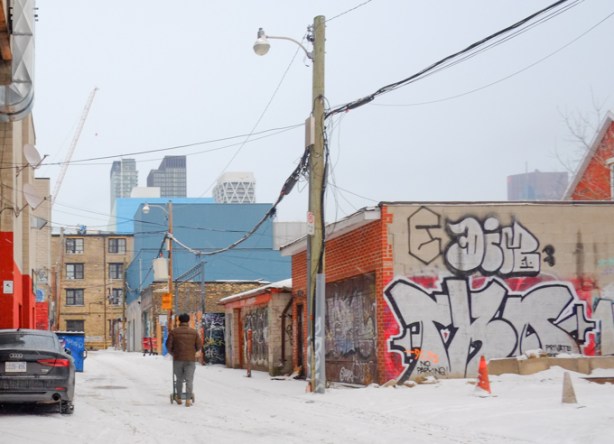 man pushing a cart along lane, in snow, with garages on his right, some with street art, utility pole with street light, tall buildings in the distance,