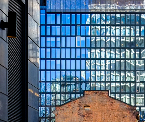 side of an old red brick smaller building in front of a wall of new condo glass and steel with many reflections in the windows