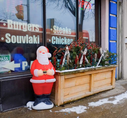 plastic santa claus stands outside a restaurant window, with words printed on window: burgers, souvlaki, steaks, chicken
