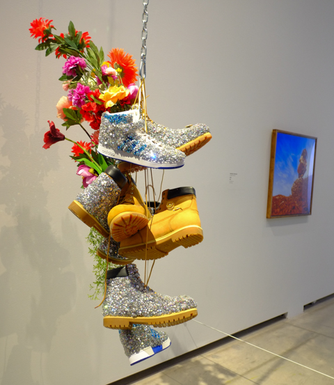 a collection of sneakers and work boots strung together on a chain and hanging from the ceiling, in an art gallery, also some artificial flowers stuck into the boots.