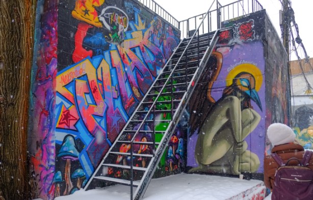 in an alley, exterior stairs to upper level balcony in alcove with two sides painted with murals, including one by sun of aquarius