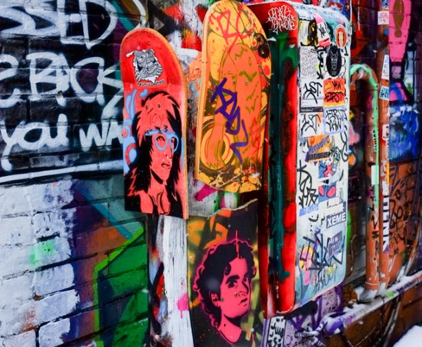 portraits painted on half skateboards, 3 different men, one in red, one in orange, and one in pink, all on the same pole in graffiti alley, work of positive creations