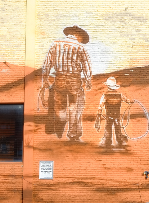 mural in brown tones, on side of wild wing restaurant, back to camera, adult male cowboy walking beside younger boy, with cowboy hats and chaps, younger one has a lasso