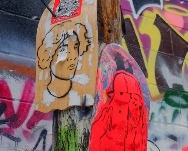 two men's portraits painted each on half of a skateboard, attached to a pole in graffiti alley. one is black line drawing on natural wood colour, and the other is very red