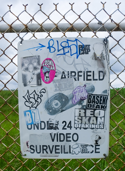sign on chainlink fence around downsview airport that says airfield, 24 hour surveillance. it is covered with graffiti stickers,