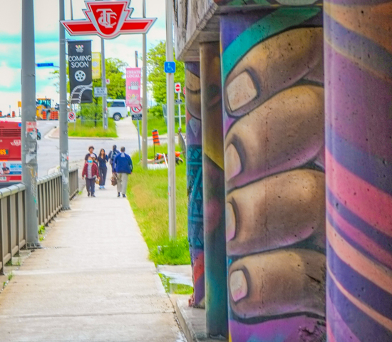 concrete pillar with street art, large brown fingers, people walking towards subway entrance