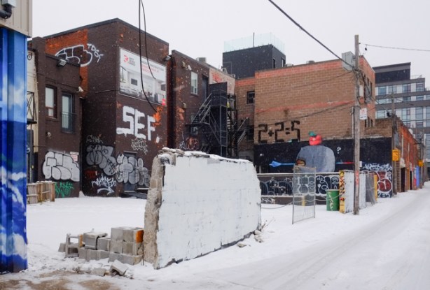 partial concrete wall, in an alley, with a row of buildings behind it, construction fence, snow, 