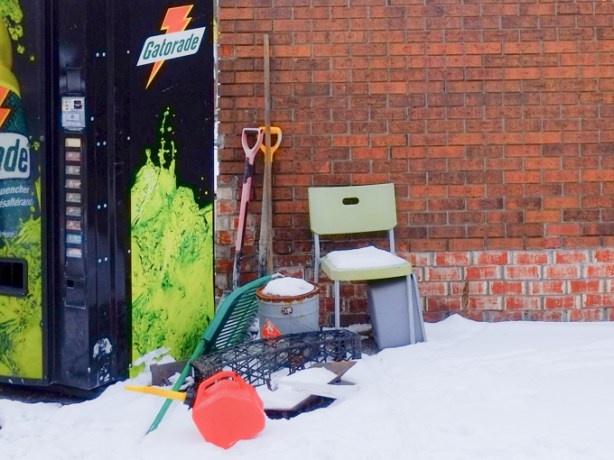 pile of stuff outside a red brick building, including a gatorade vending machine, green plastic chair, two shovels, a metal container, a red plastic gas can, and more