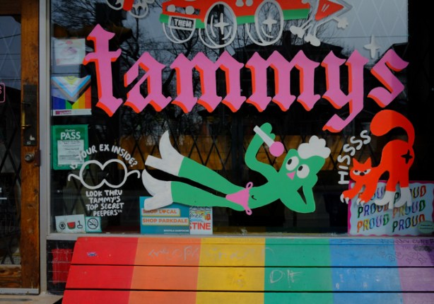 rainbow coloured bench in front of Tammys window, Gumby with white boots and pink thong, holding a microphone, 