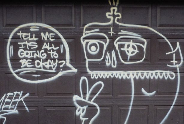 graffiti on a black garage, white drawing of a person with fingers in peace sign, with text Tell me it's all going to be okay?