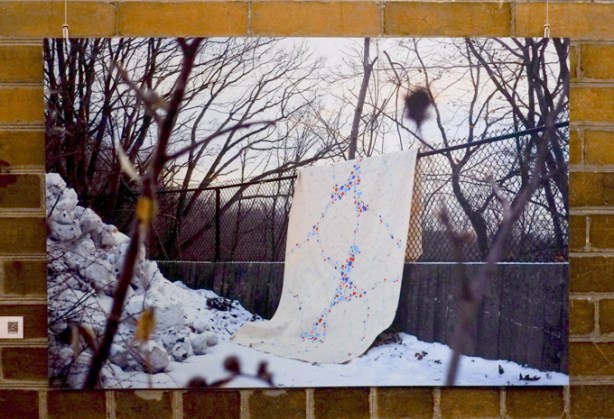 a photo of a blacket draped over oa chainlink fence, winter scene, snow on the ground, small leafless trees. 